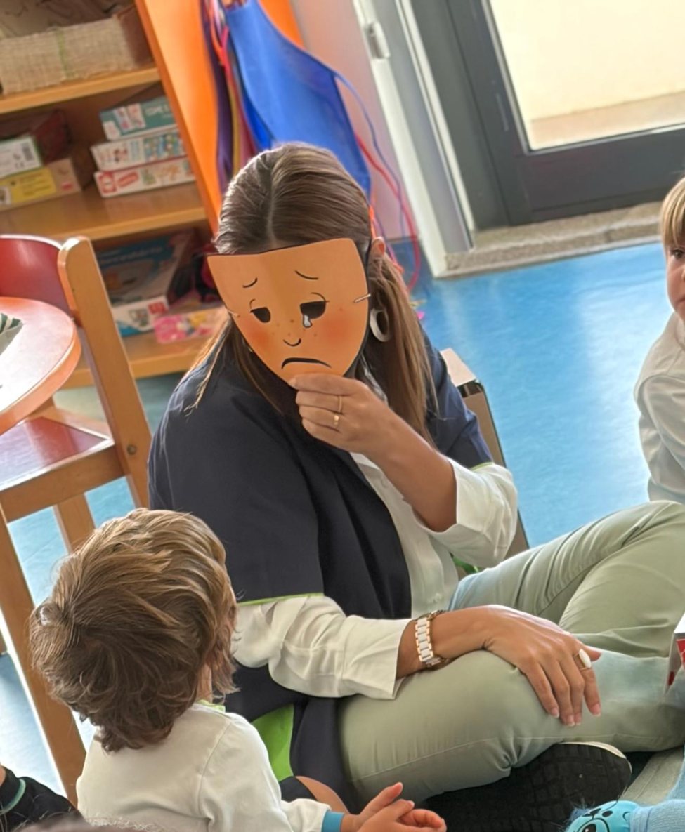 Atividade do ABC das Emoções: uma adulta com uma máscara de papel que expressa tristeza, junto de crianças sentadas em roda