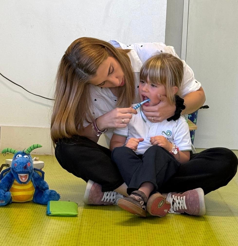 Mãe dentista a mostrar a uma criança como escovar os dentes, com uma mascote de dragão ao lado