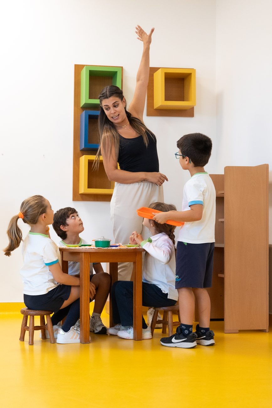 Crianças sentadas a uma pequena mesa, a brincar com a professora